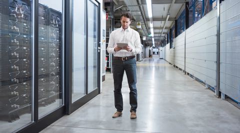 A man standing in the hallway of warehouse, looking at a tablet in his hands.